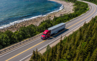 red tractor with white trailer driving along a road by the shore line and cedar trees
