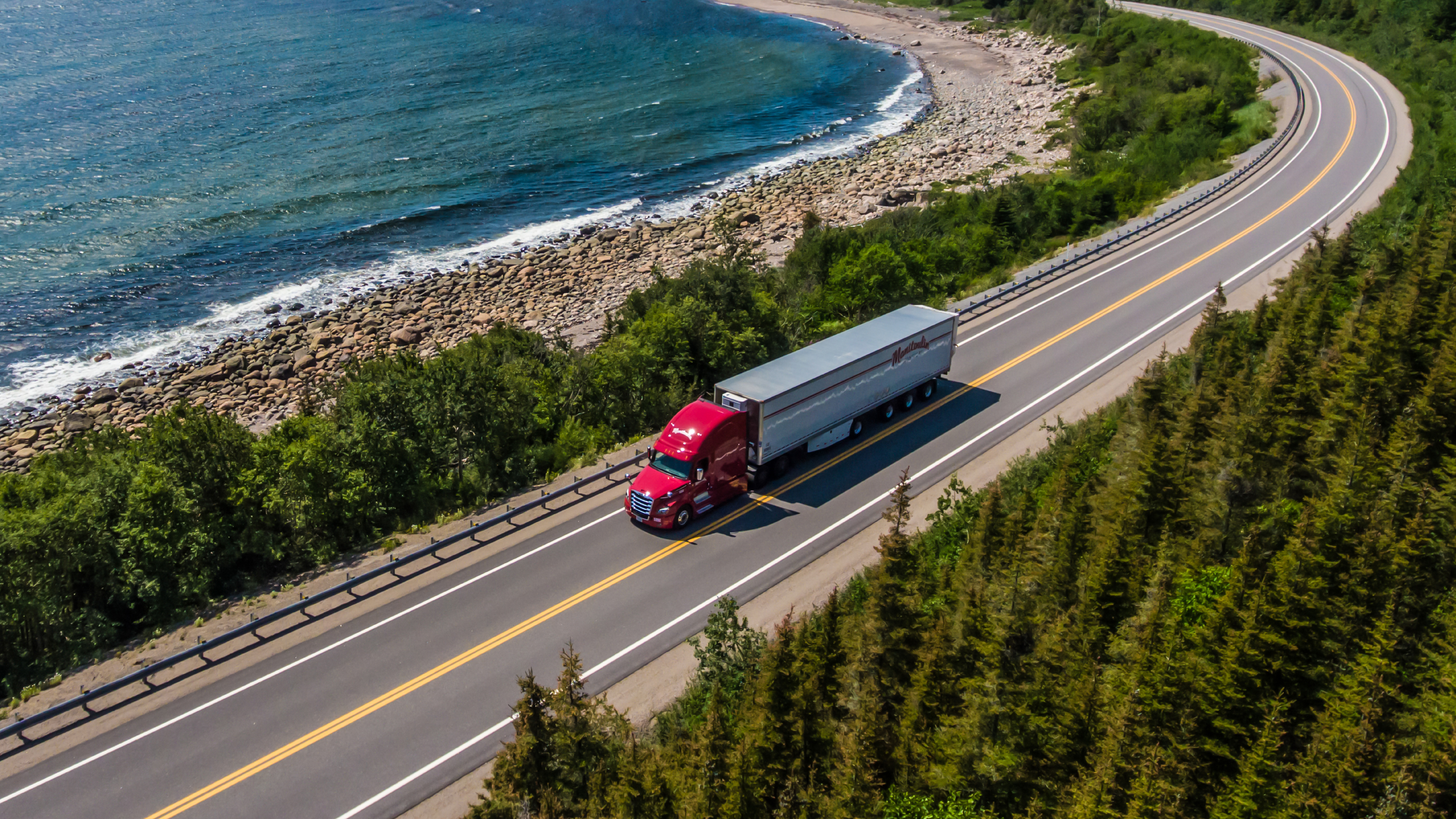 red tractor with white trailer driving along a road by the shore line and cedar trees